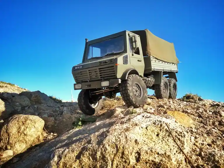 A scale model of a green military-style Mercedes-Benz Unimog truck with a canvas cover is positioned on a rocky, uneven…