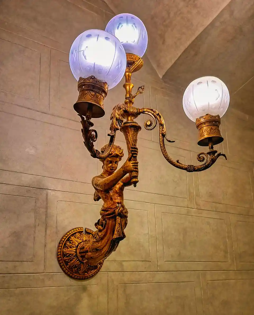 An ornate, golden wall sconce shaped like a mermaid holding a central pole with three lit globe lights, mounted on a paneled…