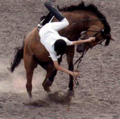 A person in a white shirt and black riding boots is thrown off a brown horse that is rearing up in a dusty arena. The rider…