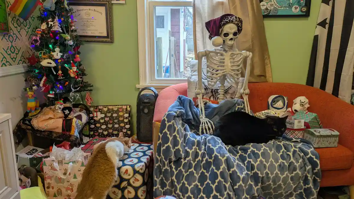 A decorated living room features a skeleton wearing a leopard-print Santa hat sitting upright on an orange couch, covered by…
