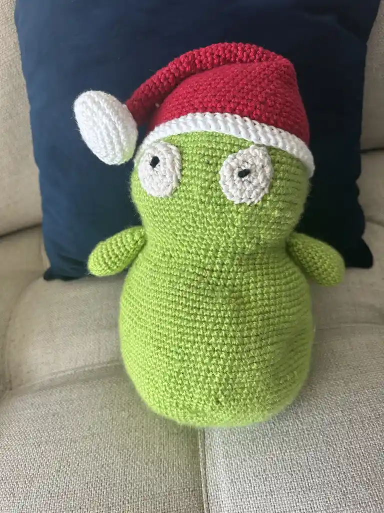 A handmade, light green crocheted plush toy resembling the Grinch, wearing a red and white Santa hat, sits on a beige couch…