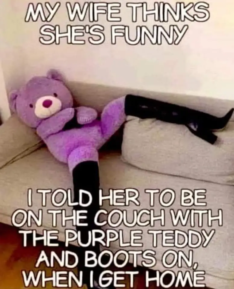 A person wearing black boots is lounging on a couch, their legs crossed, next to a large purple teddy bear. The image has…
