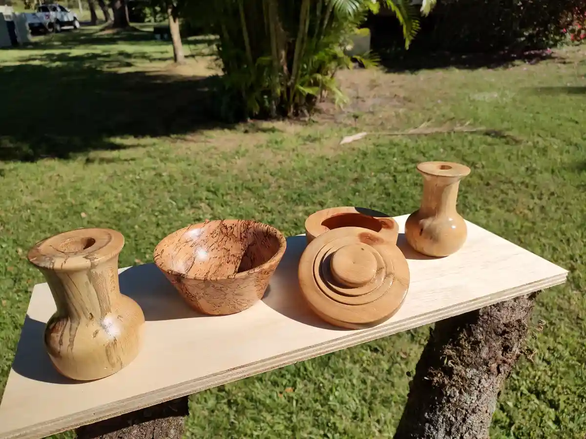 A light-colored wooden board is balanced on a tree stump outdoors, displaying four handcrafted wooden items: two small…