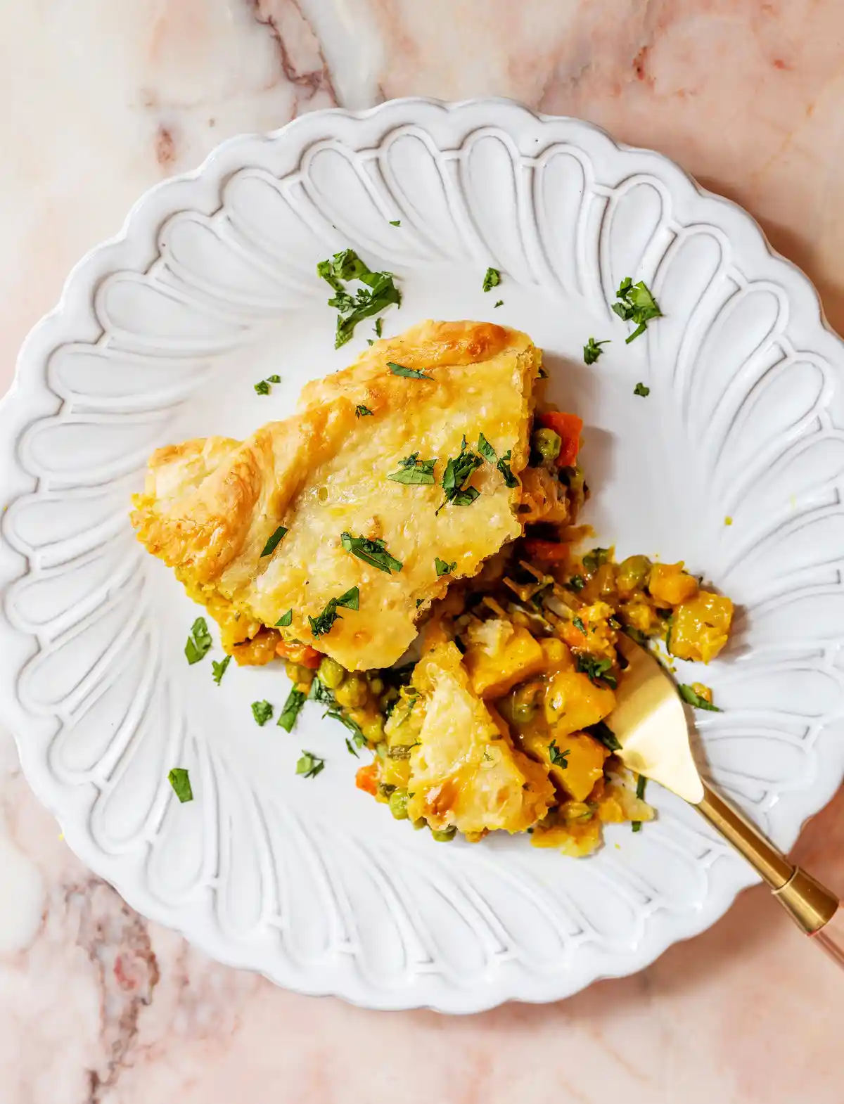 A close-up shot of a slice of savory pie on a white, scalloped plate with a gold fork, set on a pink marble surface. The pie…