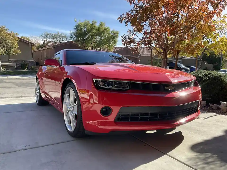 A shiny red Chevrolet Camaro is parked on a concrete driveway in a suburban neighborhood with houses and trees in the…