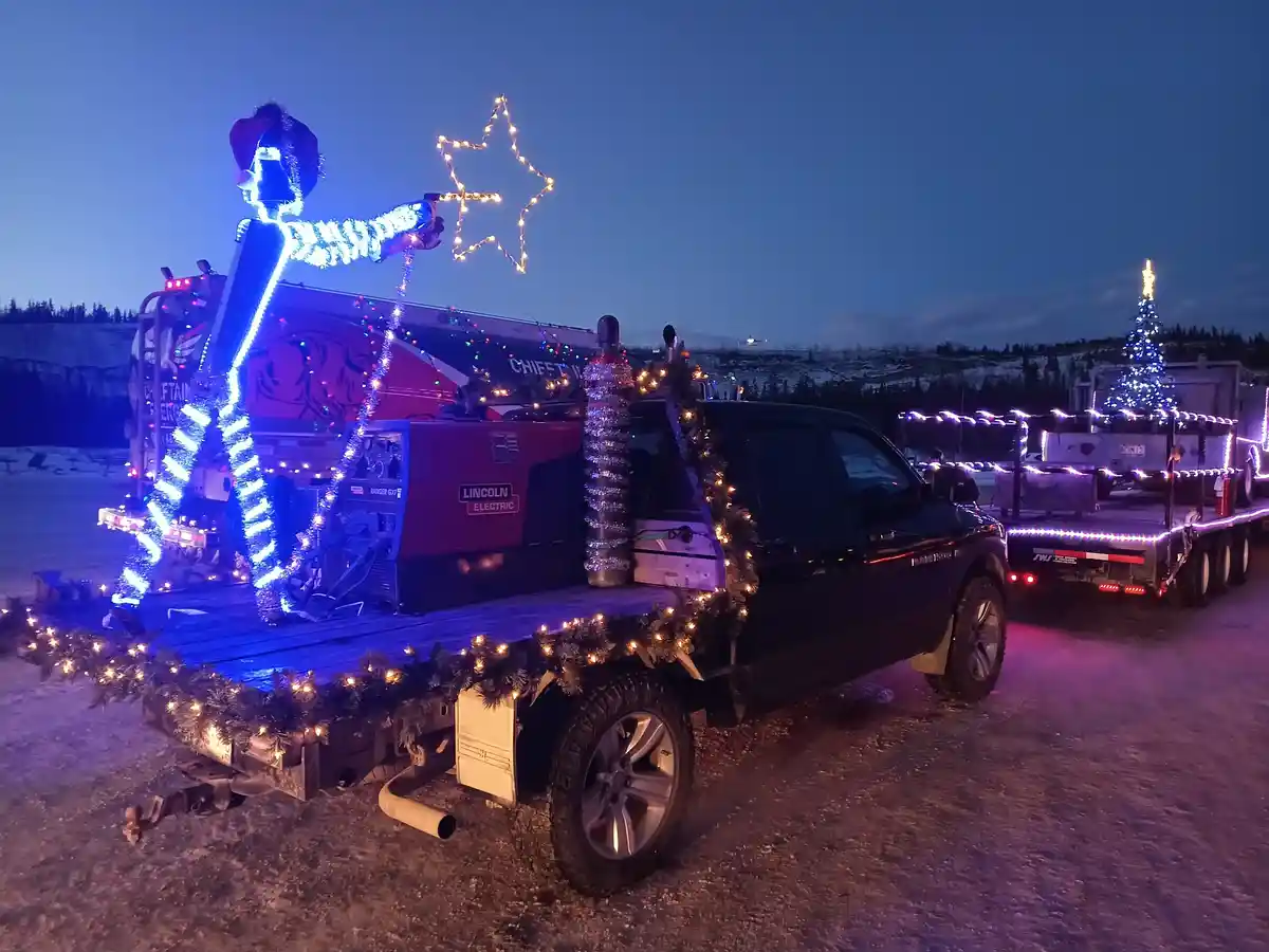 A black pickup truck decorated with Christmas lights and garlands is parked at night, with a large welding torch mounted on…
