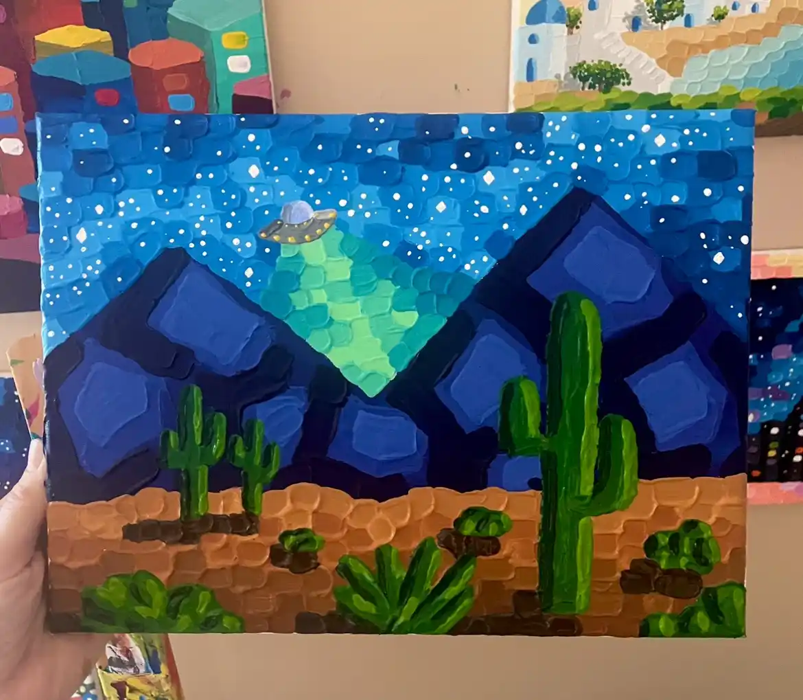 A hand holds up a textured, thickly painted canvas depicting a desert scene with cacti and mountains under a starry blue…