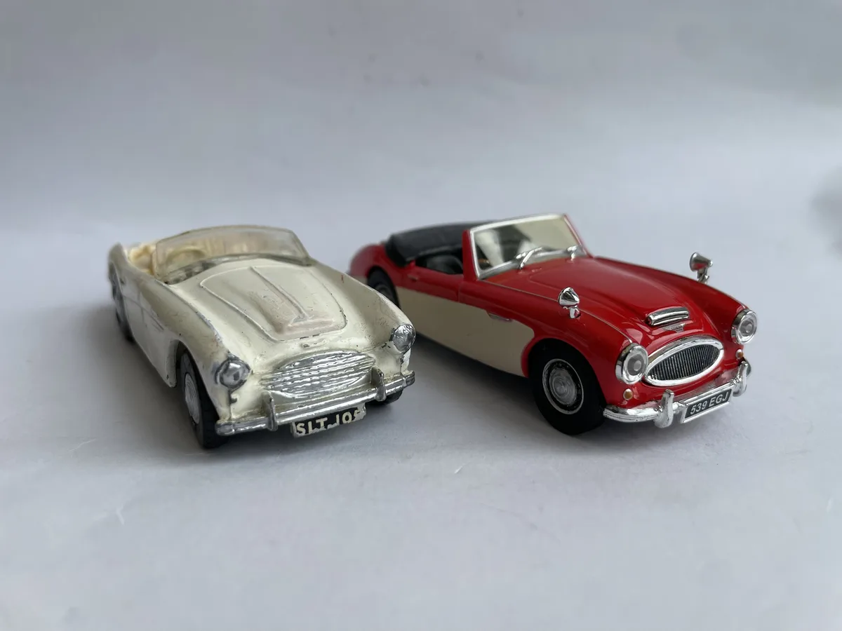 Two vintage-style toy convertibles sit side-by-side on a plain white surface; the left car is cream-colored and visibly…