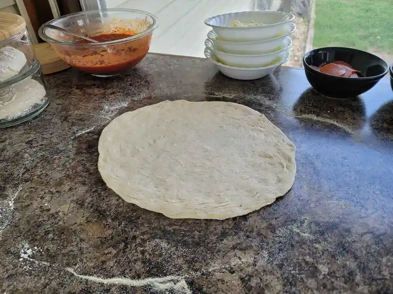 A round pizza dough sits on a flour-dusted granite countertop, surrounded by bowls of tomato sauce, shredded cheese, and…
