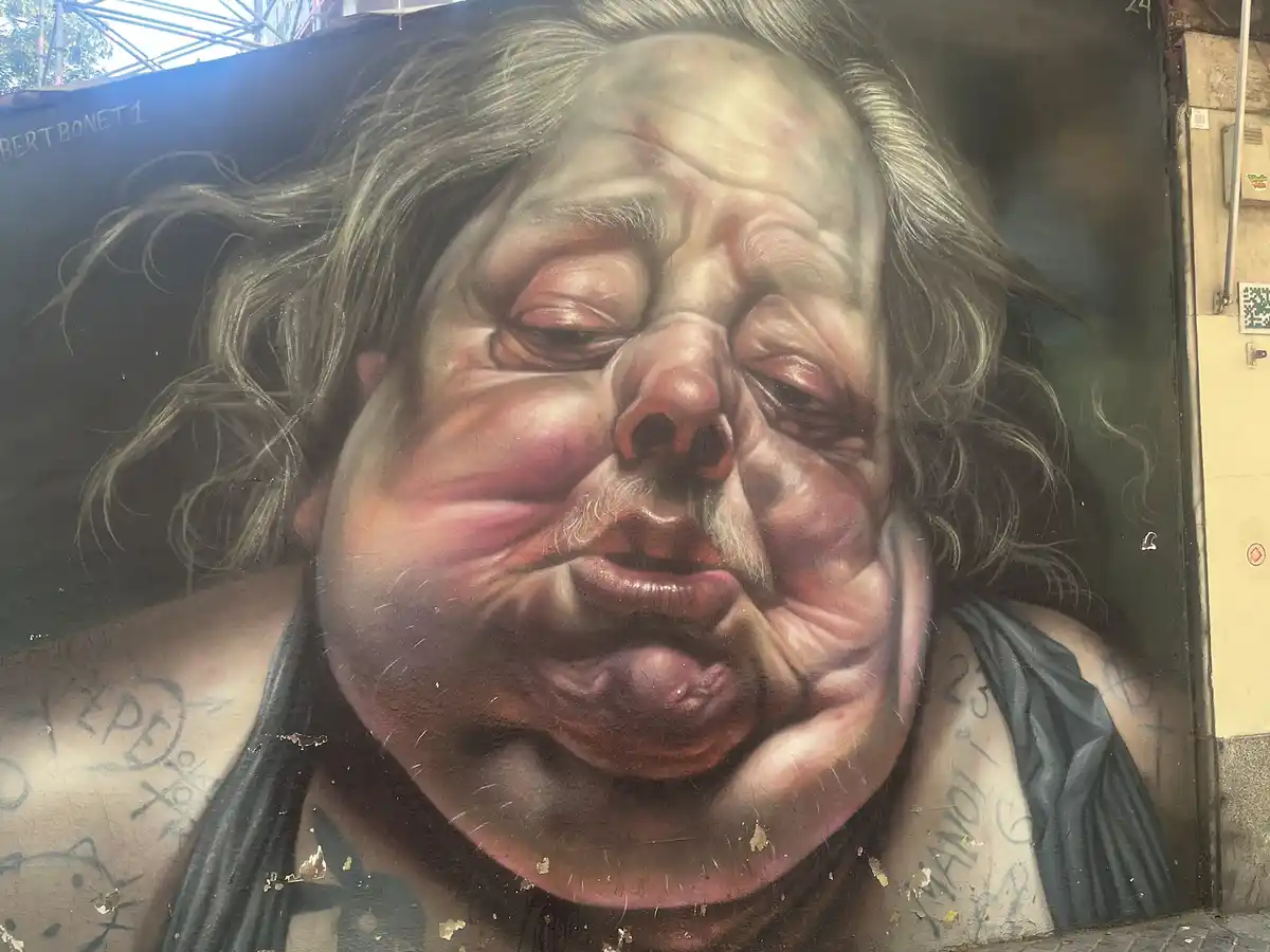A hyperrealistic street mural depicts a grotesquely obese man with closed eyes, sagging skin, and a mustache, wearing a tank…
