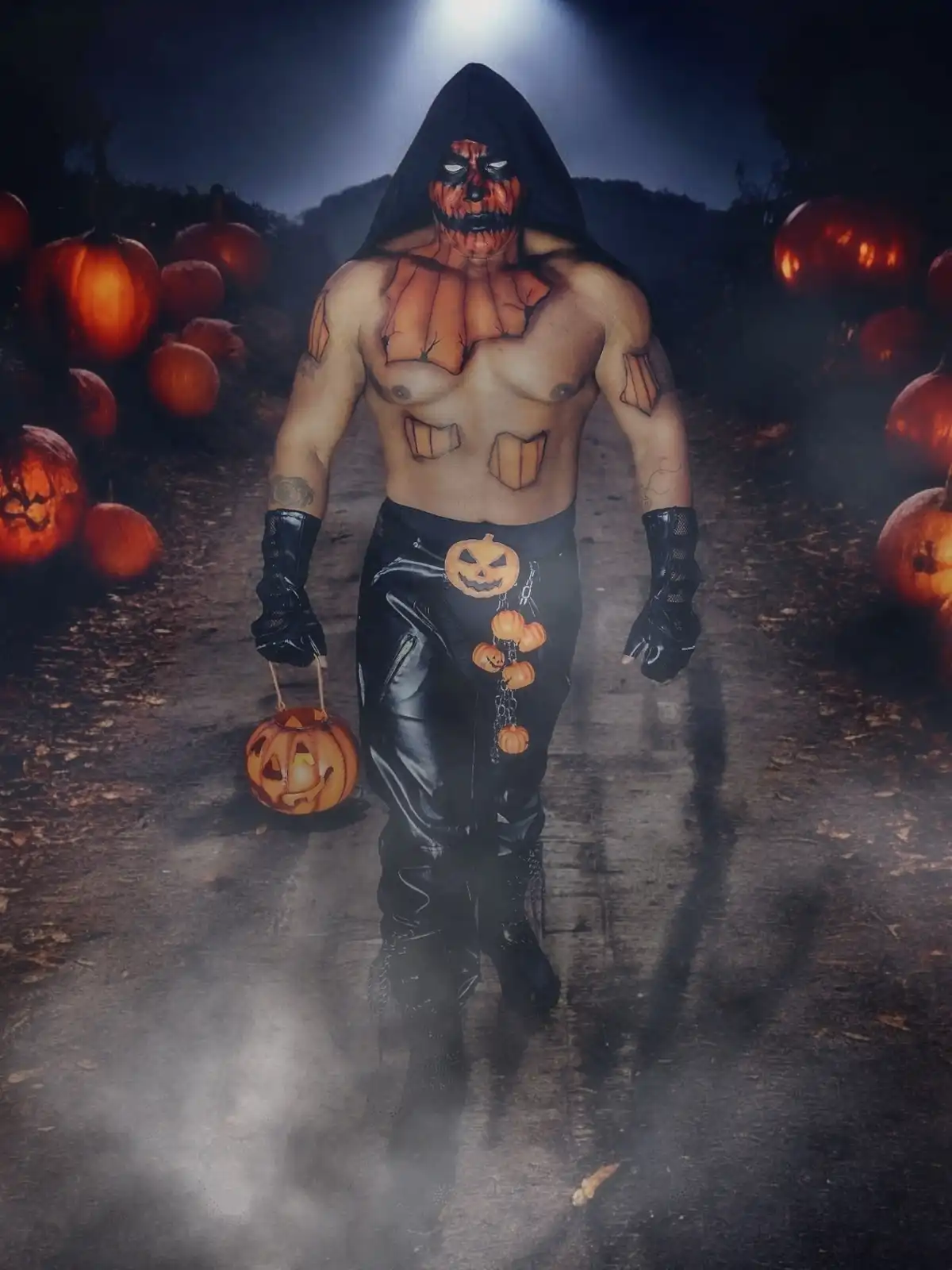 A muscular man in a black hooded outfit with pumpkin-themed body paint walks down a path surrounded by glowing pumpkins…