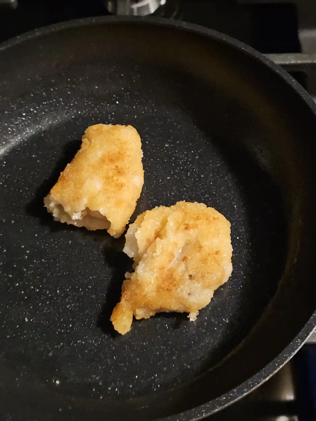 A close-up photo shows two pieces of battered and fried food, likely fish or chicken nuggets, sitting in a black speckled…