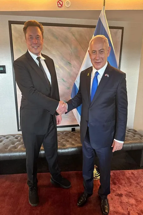Two men in suits, identified as Elon Musk and Benjamin Netanyahu, are shaking hands in front of an Israeli flag and a large…