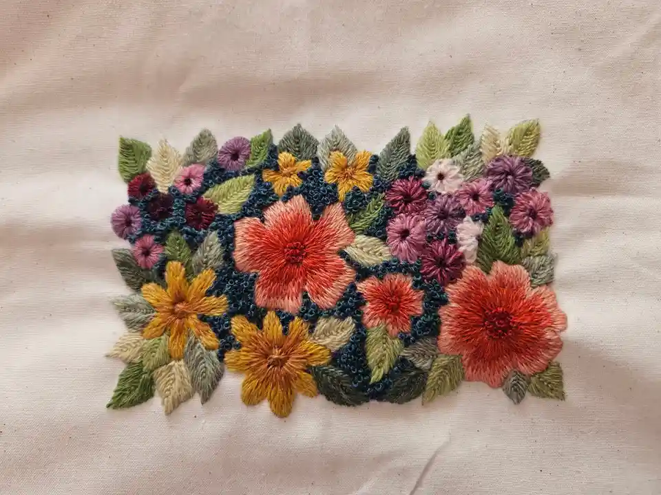 A close-up photo shows a colorful, intricate floral embroidery on a beige fabric. The embroidery features various flowers in…