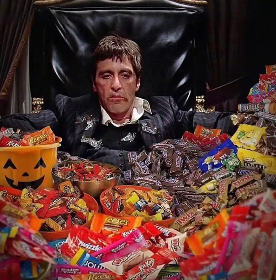 a man in a dark suit sits in a large black leather chair surrounded by piles of Halloween candy including Reese's, Twix…