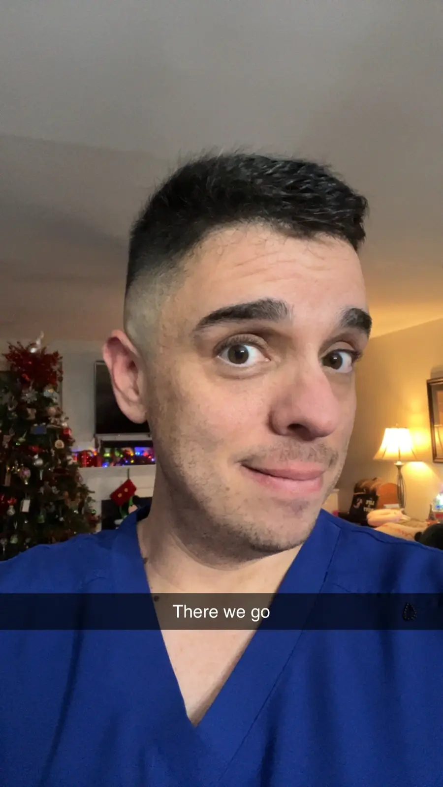 A man in blue scrubs stares directly at the camera with wide, startled eyes and a slight smirk, as if reacting to something…