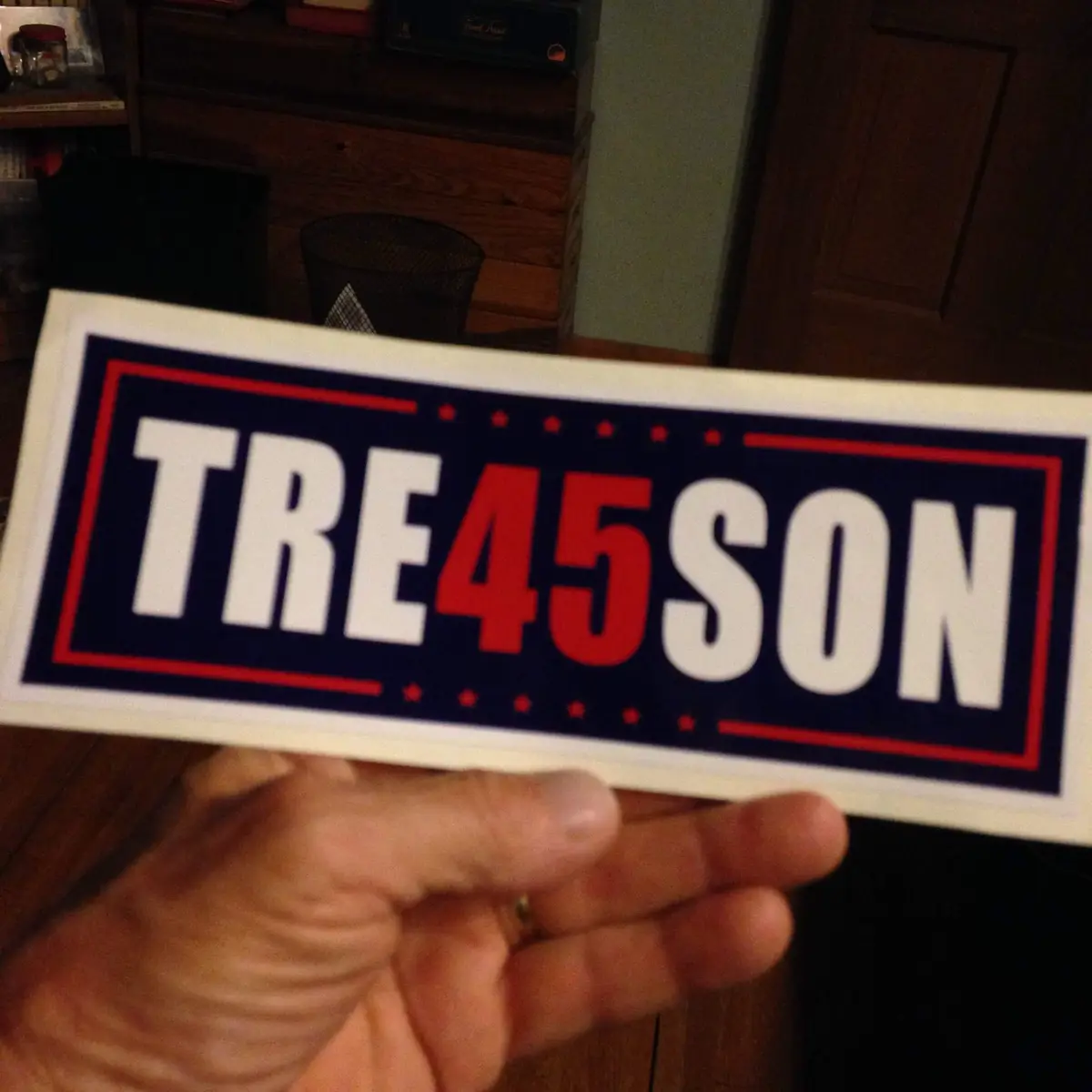 A hand holds a rectangular sticker with a blue background, red border, and white/red text that reads 'TRE45SON'. The design…