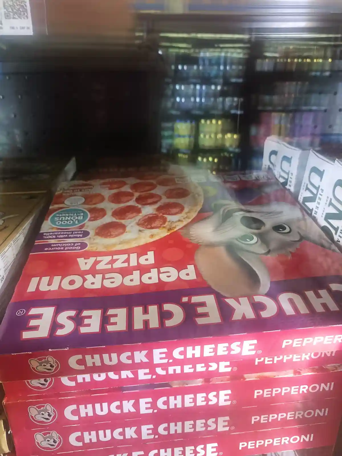 A close-up photo shows boxes of Chuck E. Cheese pepperoni pizza stacked in a refrigerated store aisle, with the brand's…