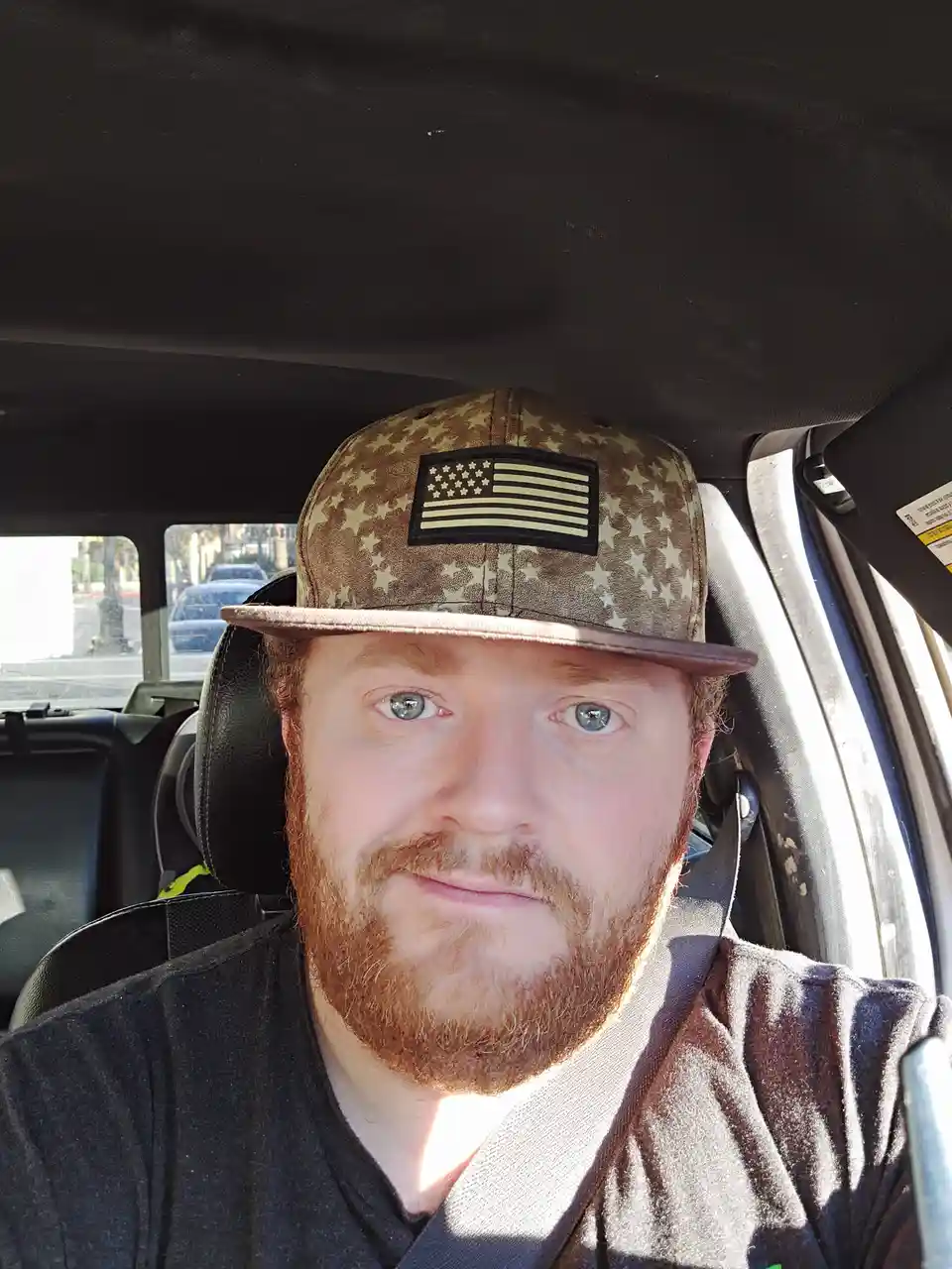 A man with a red beard and blue eyes sits in the driver's seat of a car, wearing a camouflage baseball cap with an American…