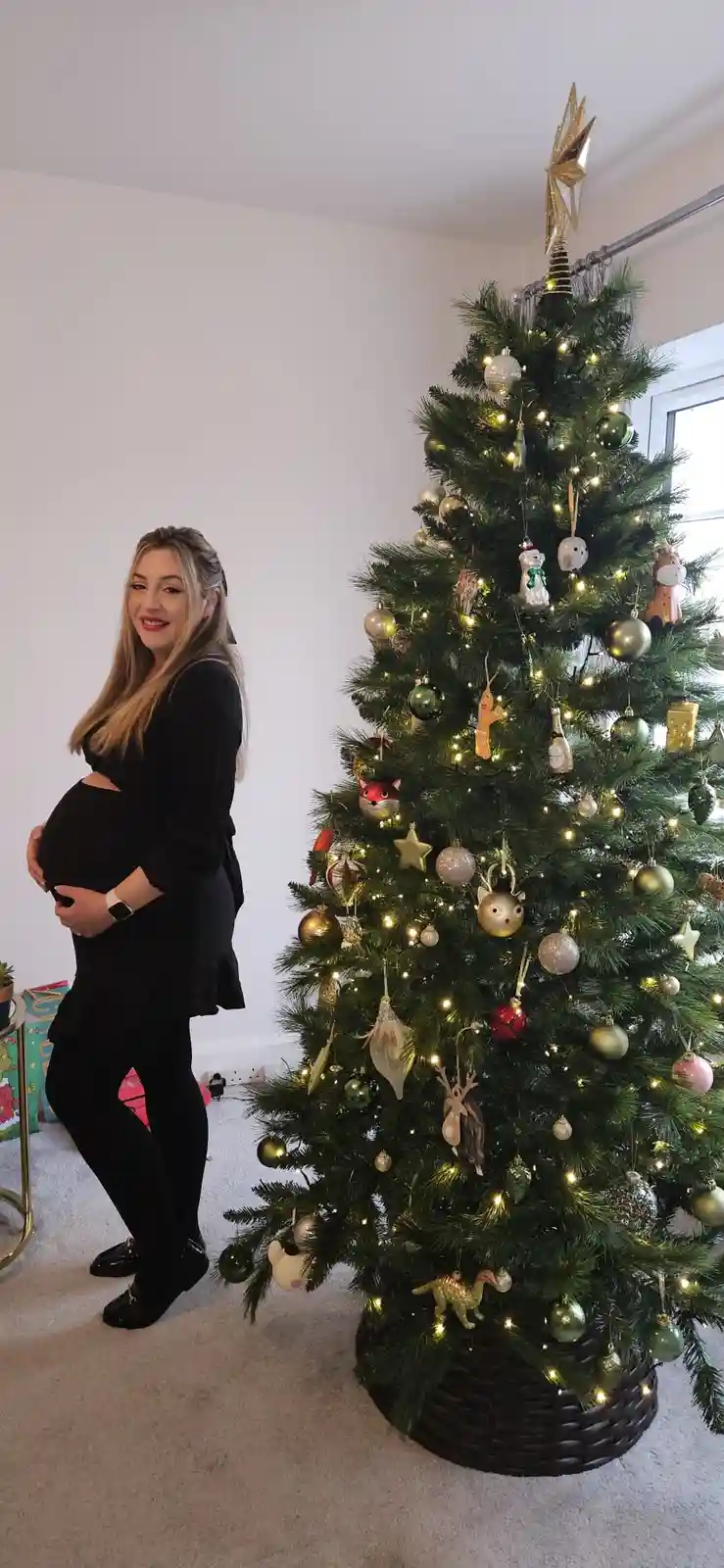 A pregnant woman in a black outfit stands beside a fully decorated Christmas tree with lights, ornaments, and a star topper…