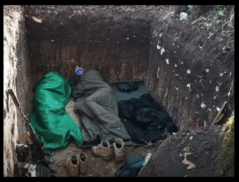 A dug-out trench or hole in the ground contains two sleeping bags, one green and one gray, with boots and a rifle nearby. A…