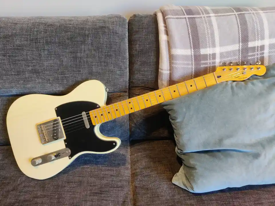 A cream-colored Squier Telecaster electric guitar rests diagonally across a gray fabric couch, its neck leaning against a…