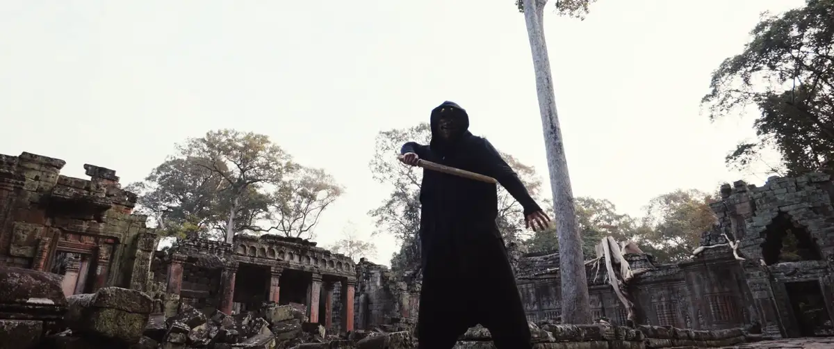 A person wearing a black hooded outfit and a grotesque, smiling mask stands in a low-angle shot amidst ancient, overgrown…