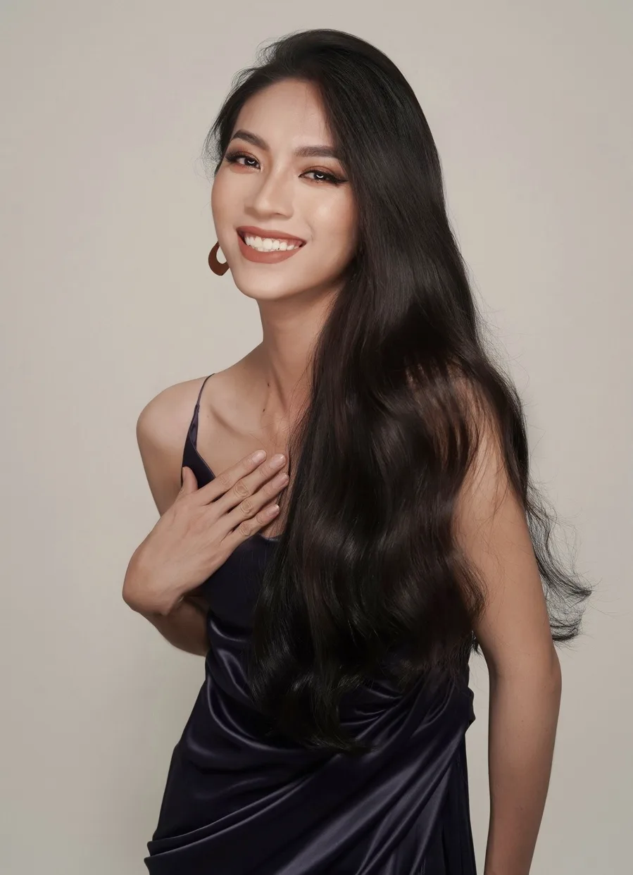 A woman with long dark hair and a warm smile poses in a dark blue satin dress, her hand gently placed over her heart against…
