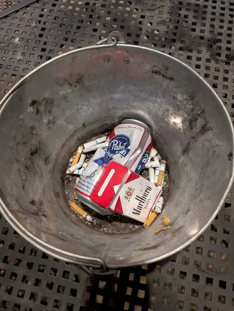 A metal bucket sits on a perforated metal table, filled with cigarette butts, a crushed Pabst Blue Ribbon beer can, and a…