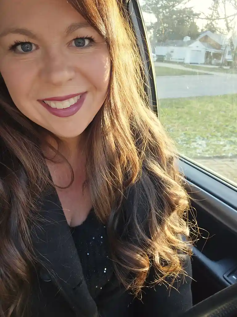 A woman with long brown hair and blue eyes smiles brightly while taking a selfie from inside a car, sunlight illuminating…