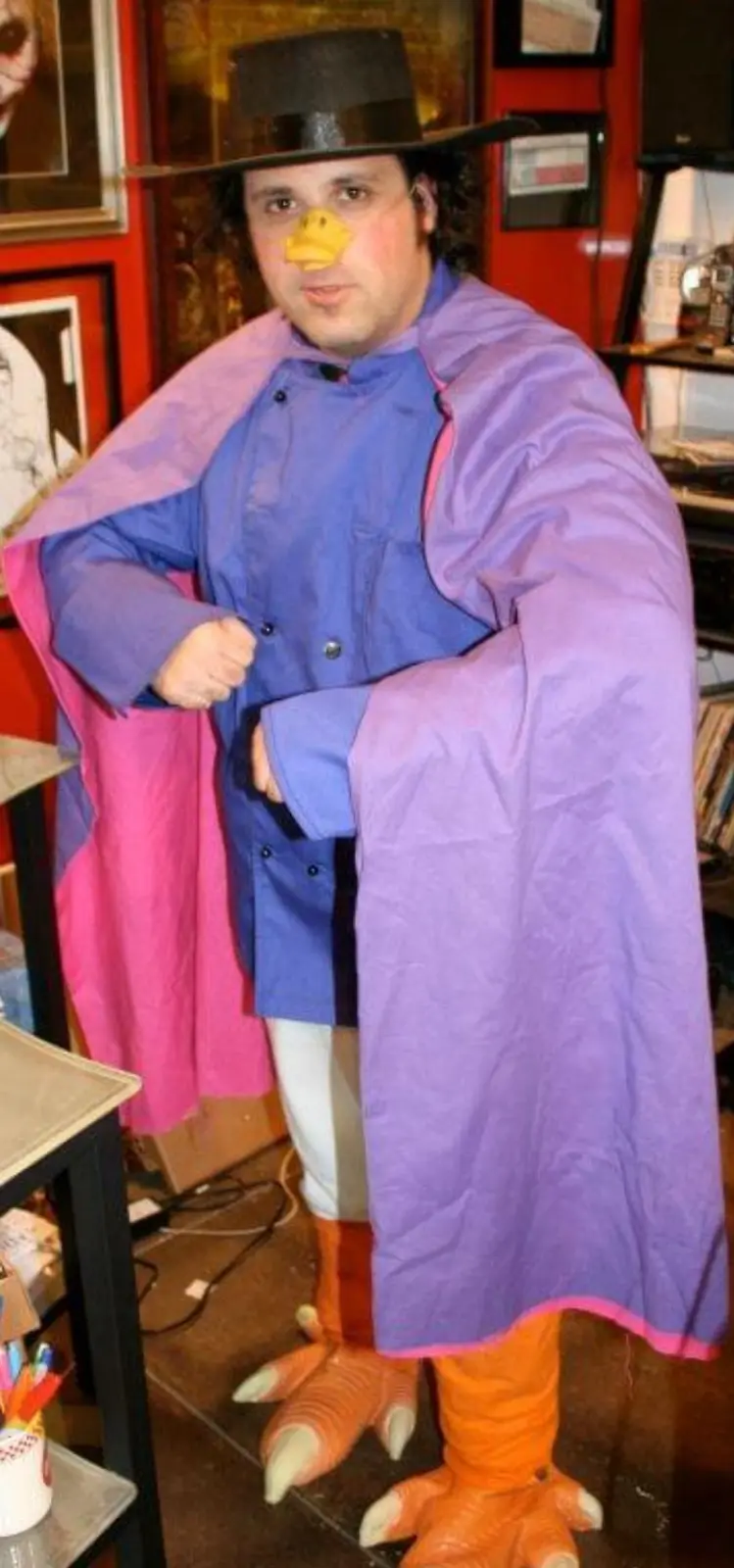 A man in a costume with a duck beak, a wide-brimmed hat, a blue coat, and a purple and pink cape, stands with orange duck…
