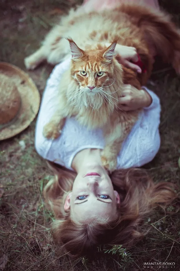 A person with long red hair lies on their back in grass, wearing a white shirt, holding a large fluffy orange cat on their…