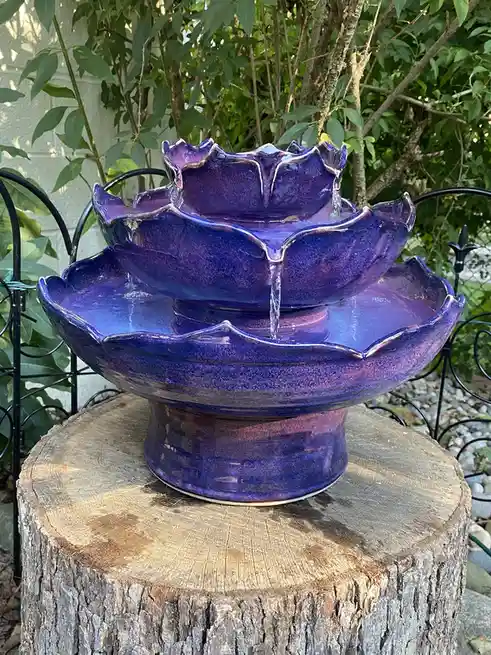 a three-tiered ceramic fountain with a glossy purple and blue glaze, shaped like overlapping lotus petals, sits on a…