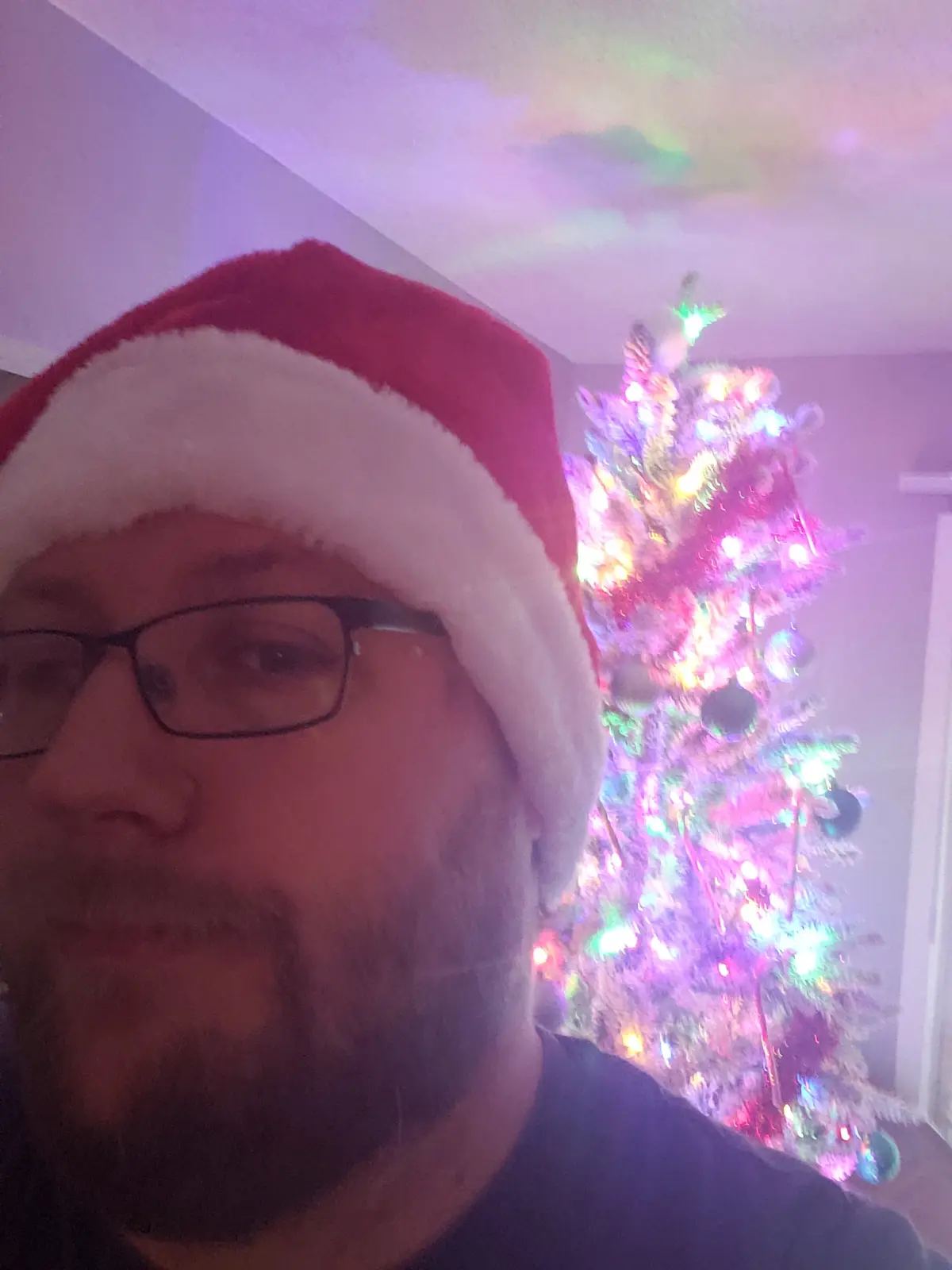 A man with glasses and a beard wears a red and white Santa hat while taking a selfie in front of a brightly lit…