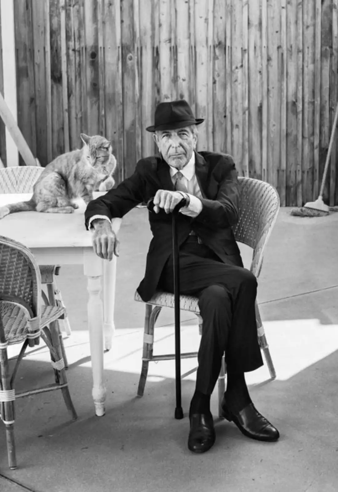 A black-and-white photo shows an elderly man in a suit and fedora sitting on a wicker chair outdoors, holding a cane. A cat…