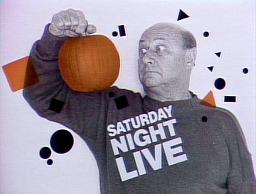 A man in a gray sweatshirt with 'SATURDAY NIGHT LIVE' printed on it holds a small orange pumpkin up with his right hand. He…