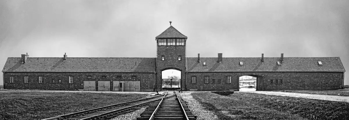 A black-and-white photograph shows the main gate of the Auschwitz concentration camp, featuring a central watchtower flanked…