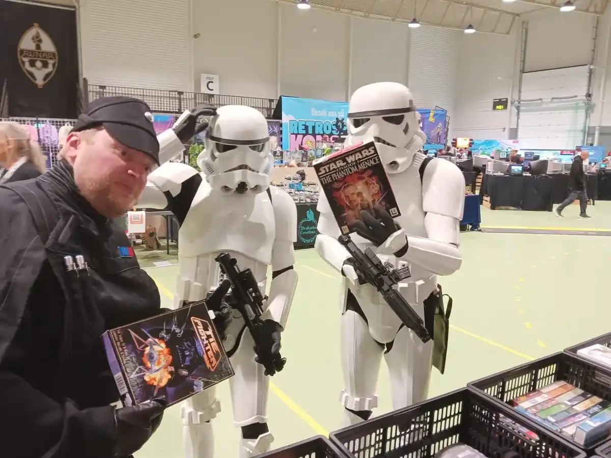 Three people at a convention: one man in a black jacket and cap holding a Star Wars game box, flanked by two Stormtroopers…