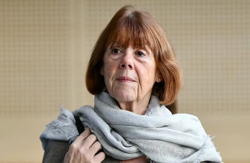 a woman with reddish-brown hair and a gray scarf looks directly at the camera with a stern, unimpressed expression. Her hand…