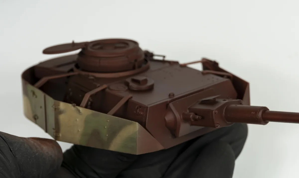 A close-up photo shows a person wearing black gloves holding a detailed model of a military tank. The tank model is mostly…