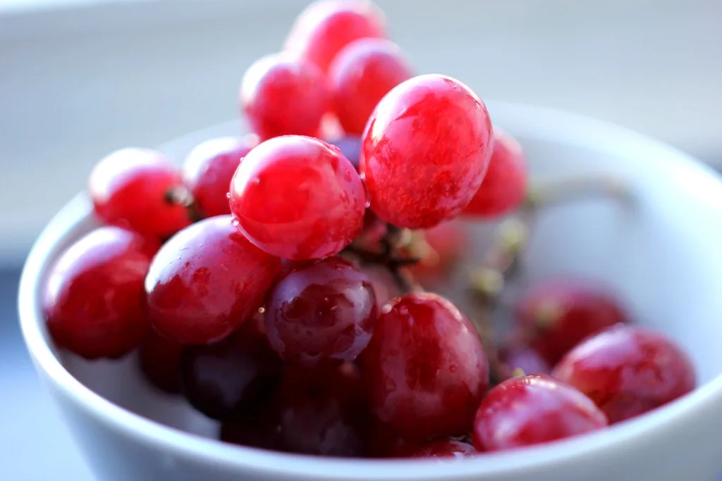 A close-up photo of a white bowl filled with shiny red grapes, some glistening with water droplets. The overlaid text reads…