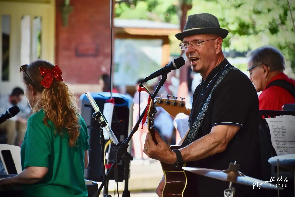 A man in a black fedora and glasses sings enthusiastically into a microphone while holding an acoustic guitar, with a woman…
