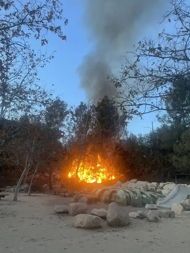 A photograph shows a large, intense wildfire burning behind a children's playground with a water feature and slide, sending…
