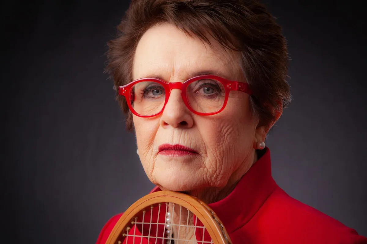 An elderly woman with short brown hair, wearing bright red glasses and a red turtleneck, holds a wooden tennis racket…