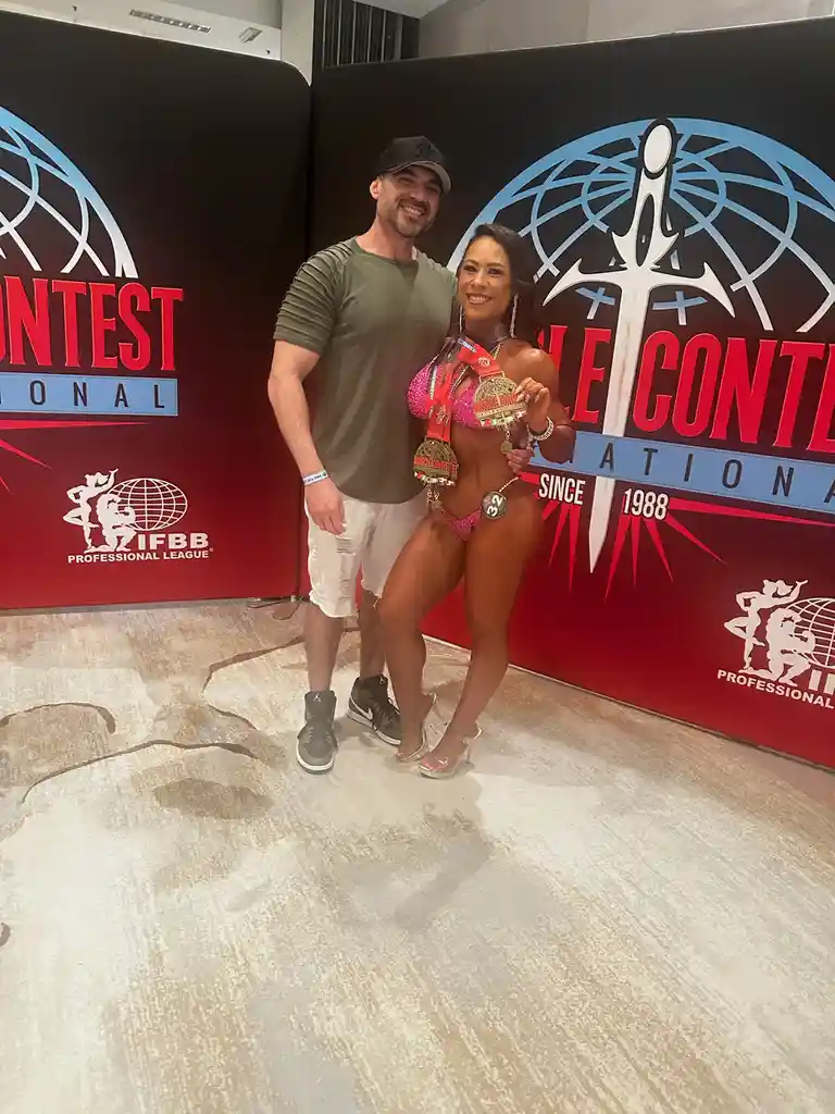 A muscular man in a green t-shirt and white shorts stands beside a smiling female bodybuilder in a pink bikini, holding…