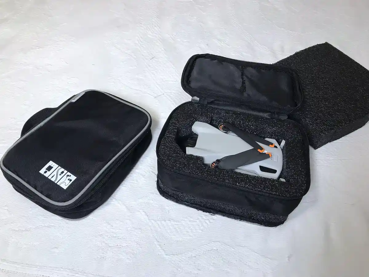 A small, folded white drone with black and orange accents sits nestled inside a black, zippered, foam-lined carrying case…
