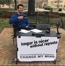 A man sits at a small outdoor desk with a microphone and papers, holding a mug. In front of him is a large white sign that…