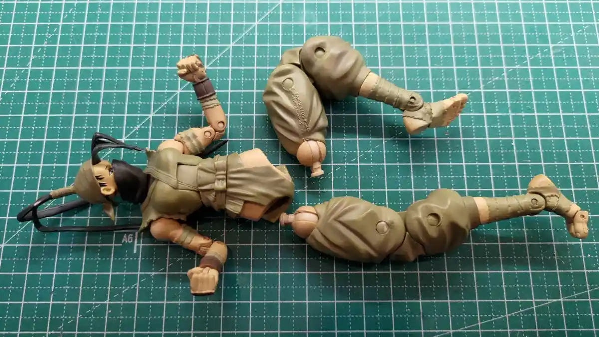 A disassembled action figure of a character with dark hair and a headband lies on a green cutting mat, with its torso, legs…
