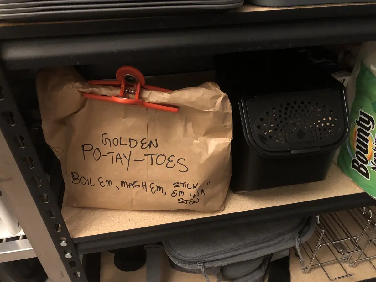 A brown paper bag on a pantry shelf is labeled in handwritten text: 'GOLDEN PO-TAY-TOES' and 'BOIL EM, MASH EM, STICK EM IN…