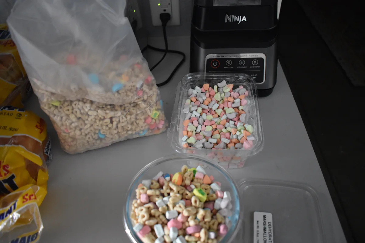A kitchen counter displays a large bag of alphabet cereal mixed with colorful marshmallows, a clear container of dehydrated…
